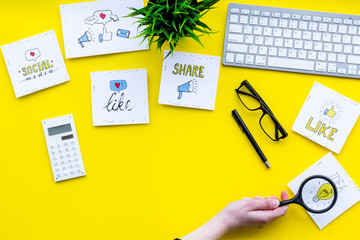 Poster - Socail media icons on work desk of marketing expert. Digital promotion of goods and services. Yellow background top view copy space. Hand with magnifier