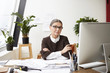 © shurkin_son - People, occupation, job and age. Confident professional senior woman architect in formal clothes and spectacles sitting at her workplace with arms folded, making drawings and using CAD program of pc