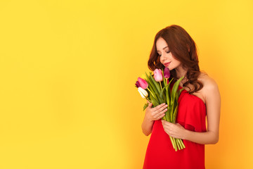Wall Mural - Young woman studio isolated on yellow in a red dress smelling tulips