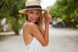© WHstudio Leushin N - Outdoor shot of pleasant looking female with tanned healthy skin, dressed in white dress and summer hat, poses in park with confident satisfied expression, likes recreation. Beautiful young woman