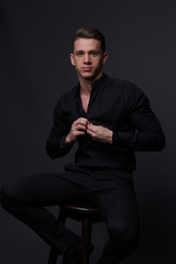 Wall Mural - A white man sits on a dark chair in a black shirt and black mills, on a gray background