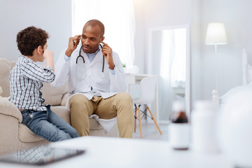 Wall Mural - Symptoms and causes. Charming nice male doctor and boy sitting on sofa while communicating and pointing at ears