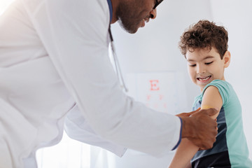 Wall Mural - Just scratch. Low angle of male doctor placing patch while pretty little boy gazing at hand and sitting