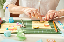 Close-up Shot Of Young Woman Decorating Handmade Greeting Card For Her Mother With Paper Flowers While Sitting At Wooden Table Of Cozy Living Room