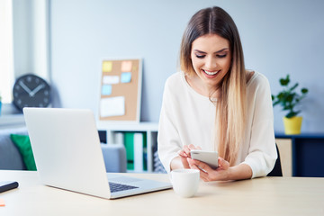 Wall Mural - Beautiful smiling woman using phone while working on laptop in home office