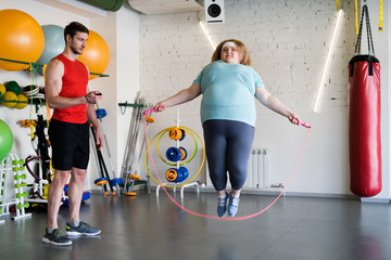 Full length portrait of overweight woman jumping with rope in gym while training with fitness instructor, copy space