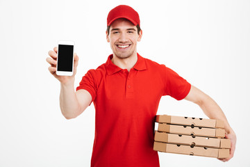 Wall Mural - Photo of happy man from delivery service in red t-shirt and cap holding stack of pizza boxes and showing smartphone, isolated over white background