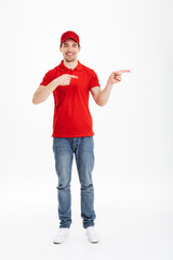 Wall Mural - Full length photo of cheerful courier in red t-shirt and cap presenting copyspace text or product with pointing fingers aside and candid smile, isolated over white background