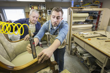 Young Caucasian Man Learning The Art Of Upholstery From A Senior Male Upholsterer.