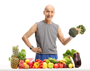 Wall Mural - Elderly man holding a broccoli dumbbell behind a table with fruit and vegetables