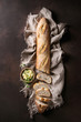 Loaf of sliced fresh baked artisan baguette bread on linen cloth with butter and herbsover dark brown texture background. Top view, copy space.