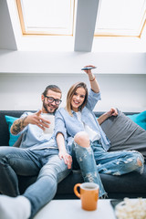 Wall Mural - Young couple sitting on the couch, drinking coffee and watching tv