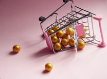 Shopping Cart. Fallen Supermarket Trolley Full Of Golden Balls On Pink Background. Consumerism Concept Photo.