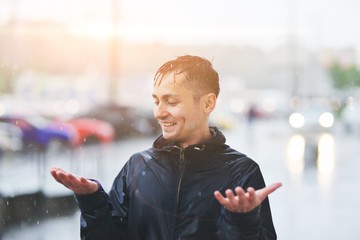 Portrait of a laughing man in a raincoat enjoing rain. He catches raindrops with his palms. Bad stormy rainy weather concept. Waterproof wear concept.