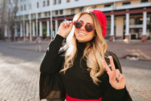 Happy Joyful Woman With Long Wavy Blond Hair Smiles At Camera Dressed Funny Sunglasses And Shows Peace Sigh On The Background Of City