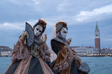 Venice Italy, February 2018. Two women in traditional costumes and masks, with decorated fans, standing in front of the Grand Canal with San Marco in the background, during the Venice Carnival