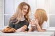 © serhiibobyk - Photo of two laughing sisters-younger and oder. Both girls have blonde straight hair. They are eating delicious sweet cookies from plate and drinking milk. Sisters sit on the white window background.