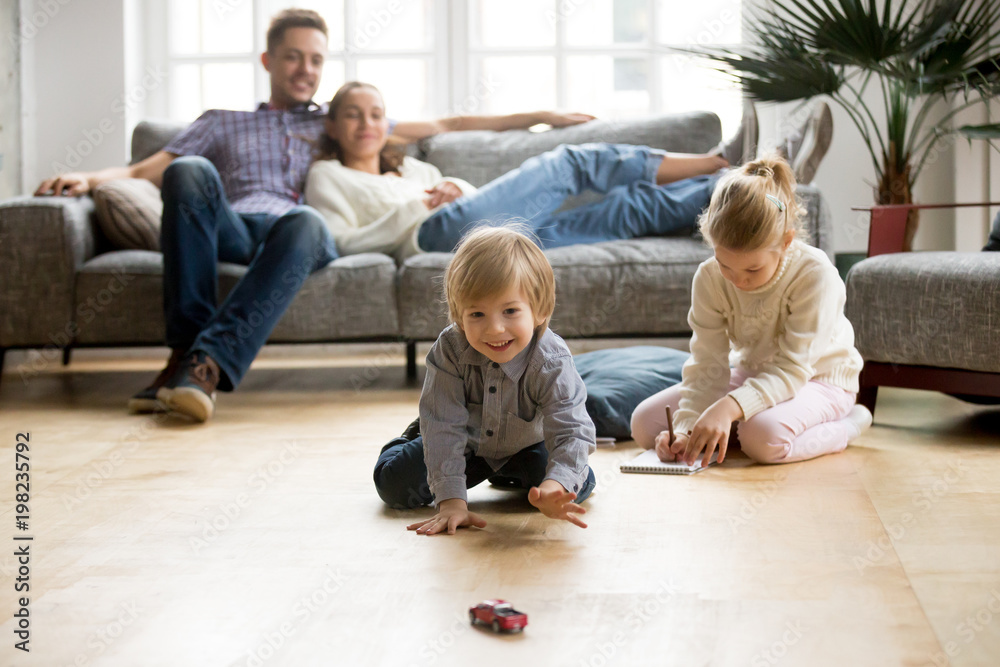 Cute kids playing while parents relaxing sofa at home together, smiling active boy entertaining with toy car near his sister on floor, happy family spending time together in living room on weekend - obrazy, fototapety, plakaty 