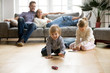© fizkes - Cute kids playing while parents relaxing sofa at home together, smiling active boy entertaining with toy car near his sister on floor, happy family spending time together in living room on weekend