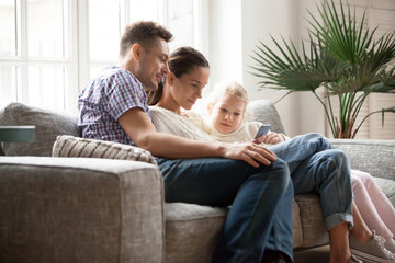 Wall Mural - Young family with daughter using app or watching educational video holding phone sitting on couch at home, married couple with little girl focused on smartphone doing mobile shopping or checking news