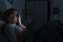 Young Sleepy Tired Woman Lying In Bed, Using Smartphone, Covering Mouth With Hand And Yawning, Eyes Closed, Can Not Sleep/Insomnia, Nomophobia, Sleep Disorder/Social Networks Addict Concept