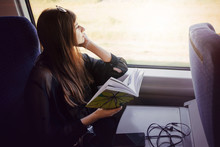 Stylish Hipster Girl Reading Book At Window Light In Train. Travelling By Train Concept. Beautiful Young Woman Holding Paper Book. Travel And Transportation. Space For Text