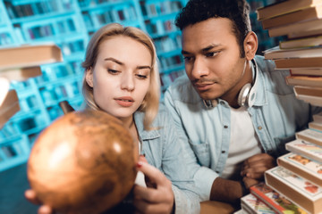 Wall Mural - Ethnic indian mixed race guy and white girl surrounded by books in library. Students are using globe.