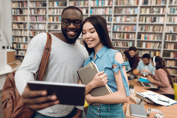 Wall Mural - Group of ethnic multicultural students in library. Black guy and asial girl are using tablet.