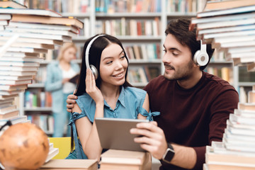 Wall Mural - Ethnic asian girl and white guy surrounded by books in library. Students are using tablet with headphones.