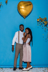 Wall Mural - stylish young african american couple posing with golden heart shaped balloon on blue