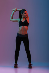 Wall Mural - Full length portrait of sportswoman in black fitness clothing looking aside while warming up and stretching her arms, isolated over colorful background