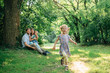 © beatleoff - Young happy family playing in the park. Little smiling girl running from her parents and brother. Man sitting with his wife and son looking at his cute daughter.