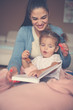 © Mladen - Cheerful mother and her little daughter at home sitting on floor and reading book together.