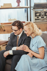 Wall Mural - happy fifties style couple looking away while sitting on sofa and holding hands