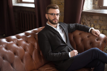 Handsome stylish confident bearded businessman in a smart suit and glasses sitting on a sofa in the office.