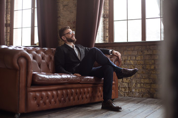 Handsome stylish confident bearded businessman in a smart suit and glasses sitting on a sofa in the office.