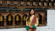 Bhutanese young woman facing the camera with a smile is wearing a kira (national dress) and standing in front of prayer wheels in monastery