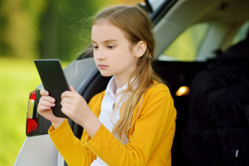 Wall Mural - Adorable little girl ready to go on vacations with her parents. Kid sitting in a car trunk and reading her ebook.