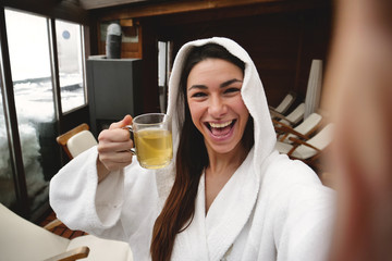  00:00 | 00:14 1×  A beautiful woman, in a wellness center, dressed in a white bathrobe, drinks a hot herbal tea and makes a video call to show the spa. Concept of: technology, relax, social network.