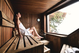 A beautiful woman wearing a white towel takes a sauna: The sauna is made of wood with a large window with a view of the snow. Concept of: relax, vacation, wellness center.