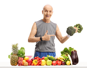 Wall Mural - Senior holding a broccoli dumbbell and pointing behind a table with fruit and vegetables