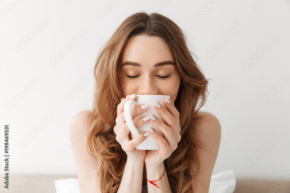 Portrait of lovely woman with beautiful brown hair drinking morning coffee or tea, while resting in bed - obrazy, fototapety, plakaty 