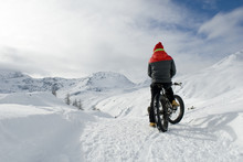 Man Use Electric Bicycle, E-bike, Ebike, Snow Covered Road, Look Mountains And Horizon, Bike With Wide Wheels To Go On Snow, Called Fatbike, Winter, Cold, Alps, Freeride, Simplon Pass, Switzerland