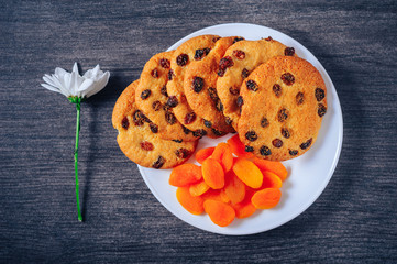 Round cookies with raisins and dried apricots on a white plate stand on a dark table with a white flower, top view