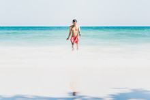 Young Man On Beautiful Sunny White Sand Beach On Ko Samet, Thailand: Asian Man In Red Swimming Trunks Walks From Turquoise Sea Waters To The Wide Empty Shore, Happy & Wet After Swim. Wide Copy Space