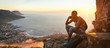 Panoramic image of a young black man sitting on the top of a mountain talking on a mobile phone during an amazing sunset, with a beautiful view of the ocean and cityscape in the background.