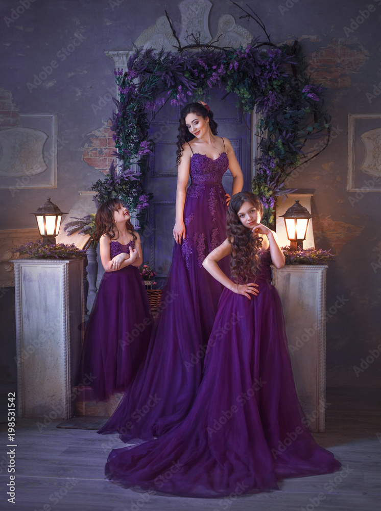 A family outfit. Mom and two daughters posing against the backdrop of a blossoming courtyard in sumptuous purple-burgundy dresses with a very long train. Girls have long curly hair. Art photo Stock A family outfit. Mom and two daughters posing against the backdrop of a blossoming courtyard in sumptuous purple-burgundy dresses with a very long train. Girls have long curly hair. Art photo Stock