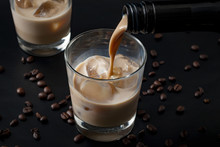Pouring irish cream in a glass with ice, surrounded by coffee beans on a dark black background