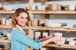 © LIGHTFIELD STUDIOS - beautiful smiling potter with ceramic dishware on shelves in workshop