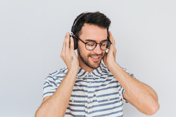 Sticker - Horizontal shot of handsome happy young male wearing striped shirt and trendy glasses, with headphones on head, listening favorite music, isolated on white studio background.Copy space for advertising